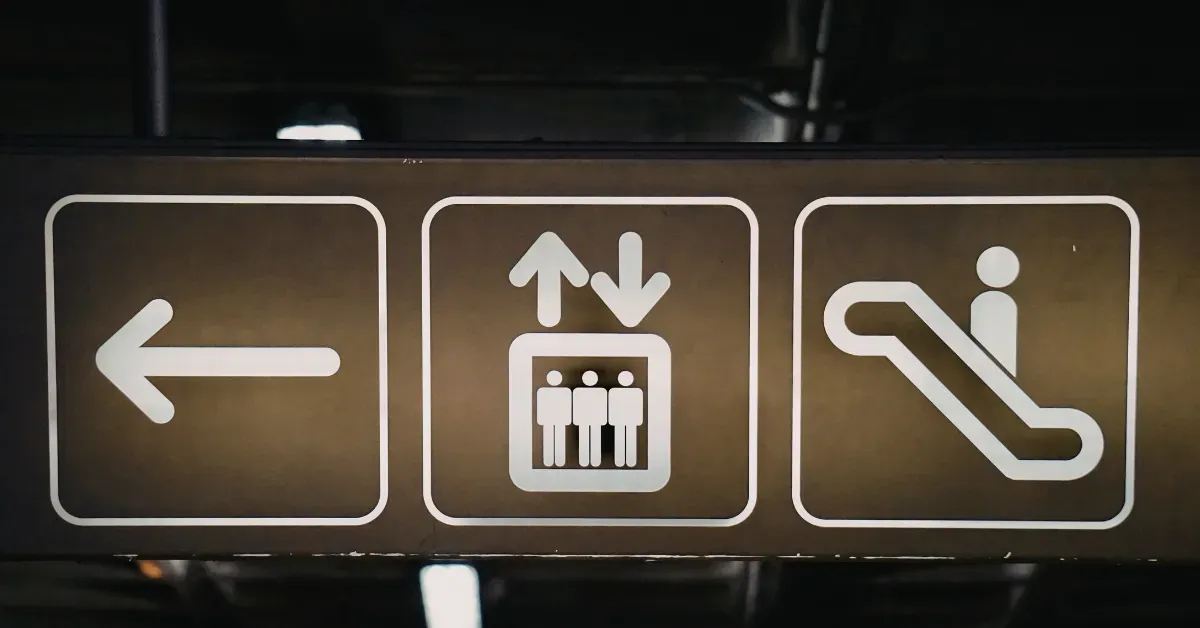 Indoor directional signage featuring white icons for an arrow, elevator, and escalator on a dark background for clear wayfinding.