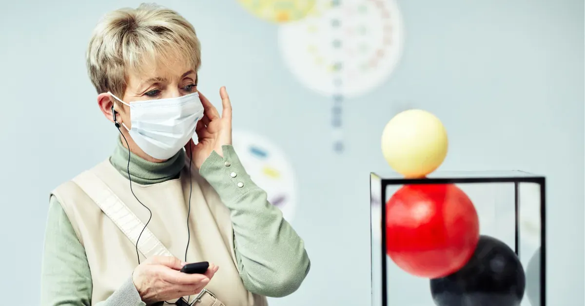Older woman wearing a face mask using an audio guide in a museum with colorful spherical art in the background.