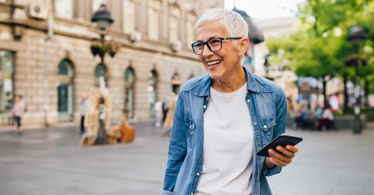 Senior woman with white hair wearing glasses and denim shirt using a smartphone to follow a heritage walk app outdoors.