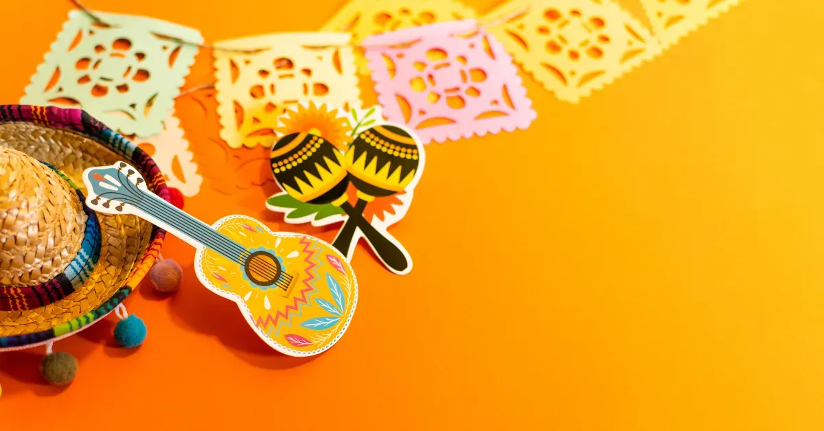 A festive display of a mini sombrero, maracas, and guitar cutouts with colorful papel picado banners on an orange background.