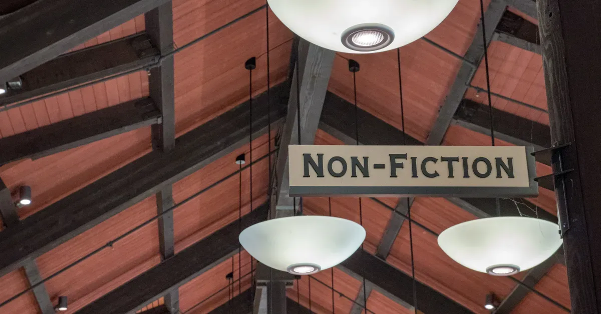 A hanging identification sign for a 'NON-FICTION' section in a library or bookstore, featuring bold black lettering against a wooden ceiling structure.