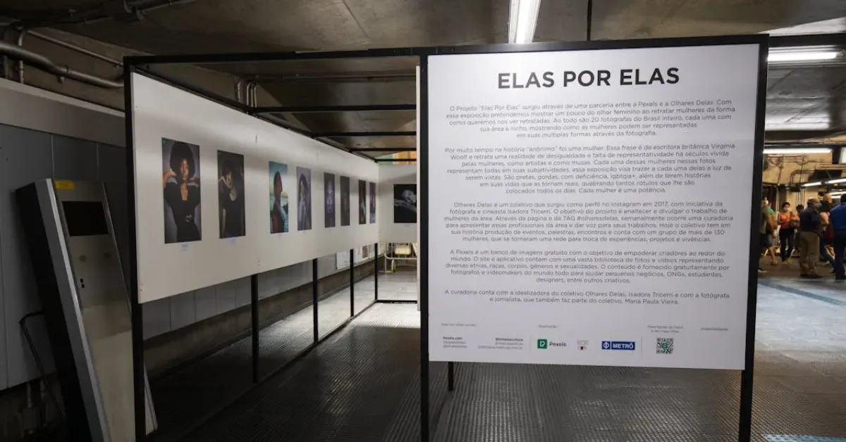 Large white informational signage board titled 'ELAS POR ELAS' providing exhibition details next to a gallery of portraits in a public transit station.