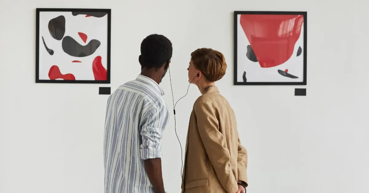 Two museum visitors using an audio guide device while viewing paintings in a gallery.