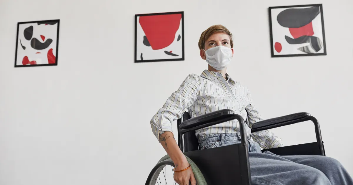 A woman in a wheelchair wearing a face mask while viewing abstract art paintings in a bright, modern museum gallery.