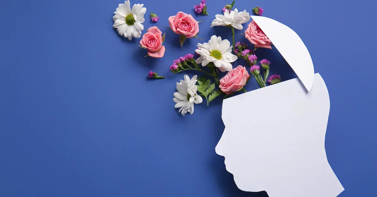 A paper cutout of a human profile with colorful flowers blooming out of the head against a solid blue background.