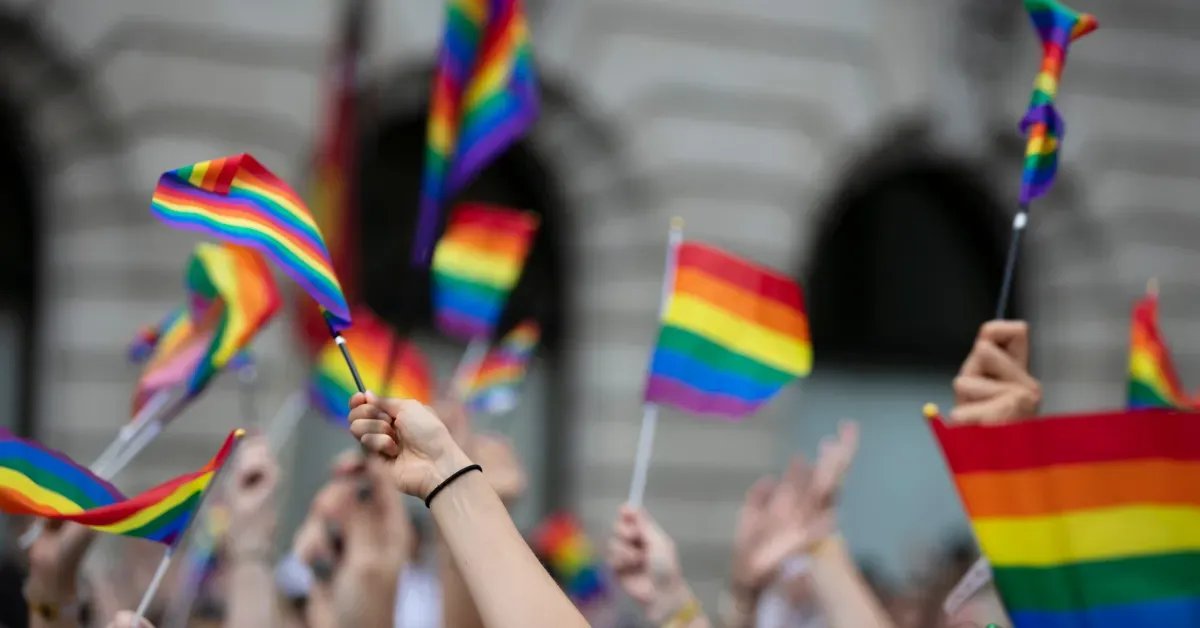 A crowd of people holding and waving small LGBTQ+ rainbow flags in front of a grey stone building during a parade.