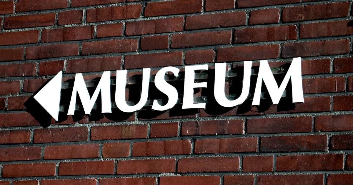A white 'MUSEUM' directional sign with an arrow mounted on a red brick wall, showcasing clear outdoor wayfinding signage.